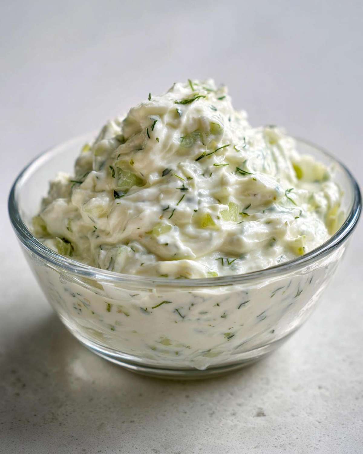A close-up of a creamy, chunky pickle dip garnished with fresh dill in a clear glass bowl.