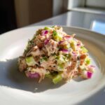 A mound of creamy salmon salad featuring flaked salmon, diced red onion, and celery on a white plate.