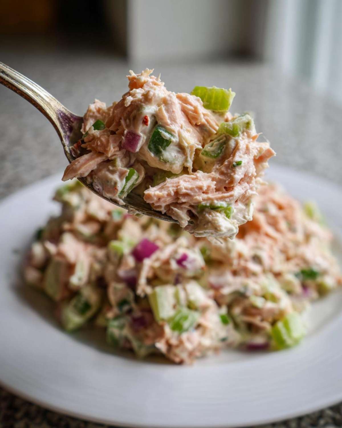 A spoonful of creamy salmon salad, featuring flaked salmon, celery, and red onion, lifted above a plate.