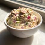 A close-up of a white bowl filled with creamy tuna dip mixed with chopped celery and red onion.