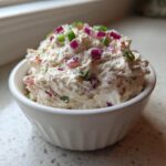 A close-up of creamy tuna dip served in a small white bowl, topped with diced red onion and green chives.