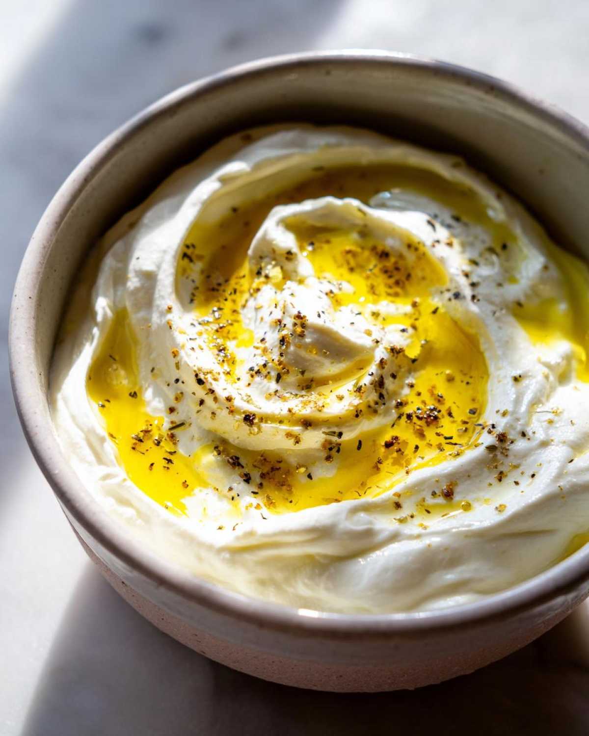 Close-up of creamy whipped feta dip swirled in a bowl, topped with golden olive oil and herbs.