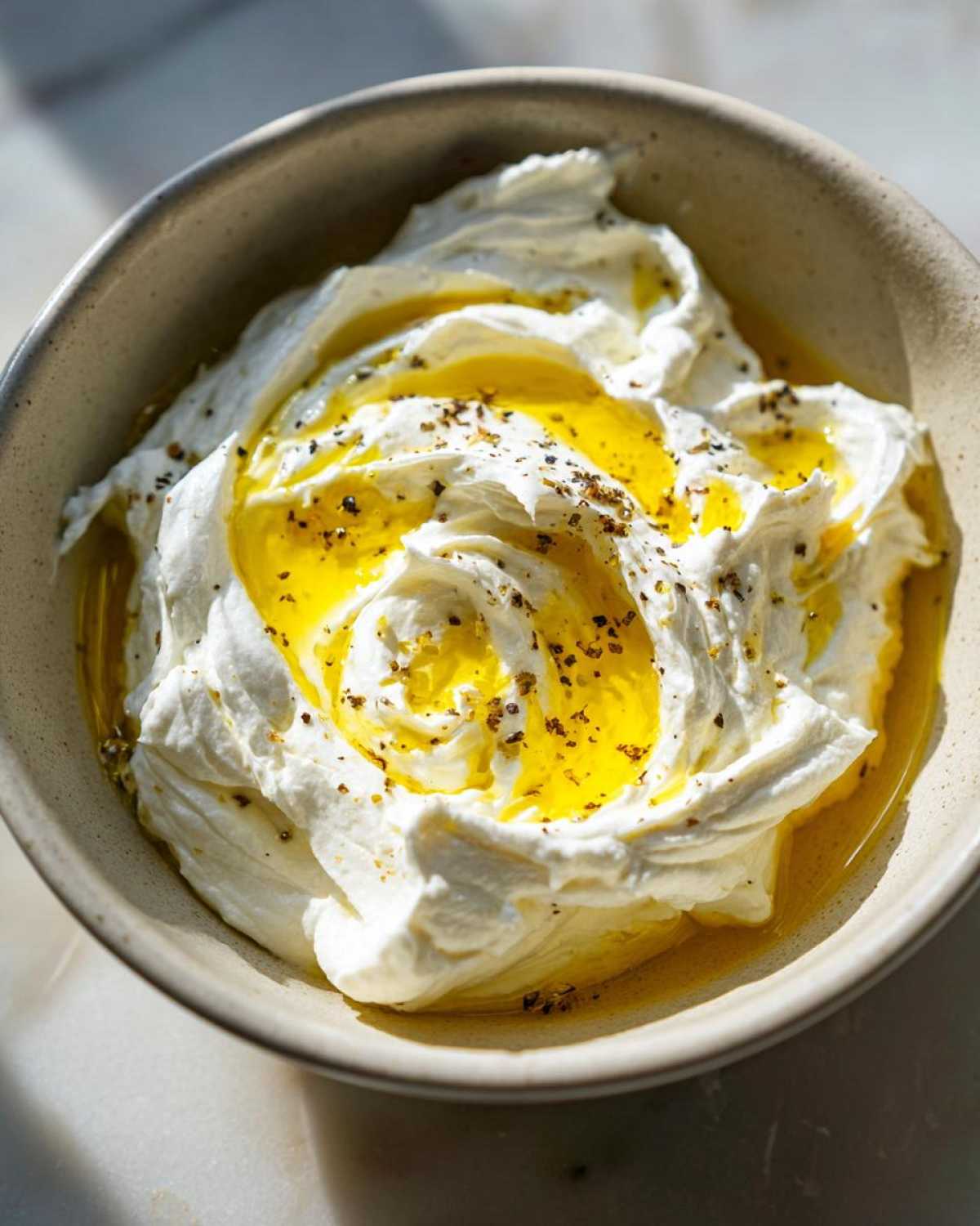 A close-up of creamy whipped feta dip swirled high in a bowl, drizzled generously with bright yellow olive oil and cracked pepper.