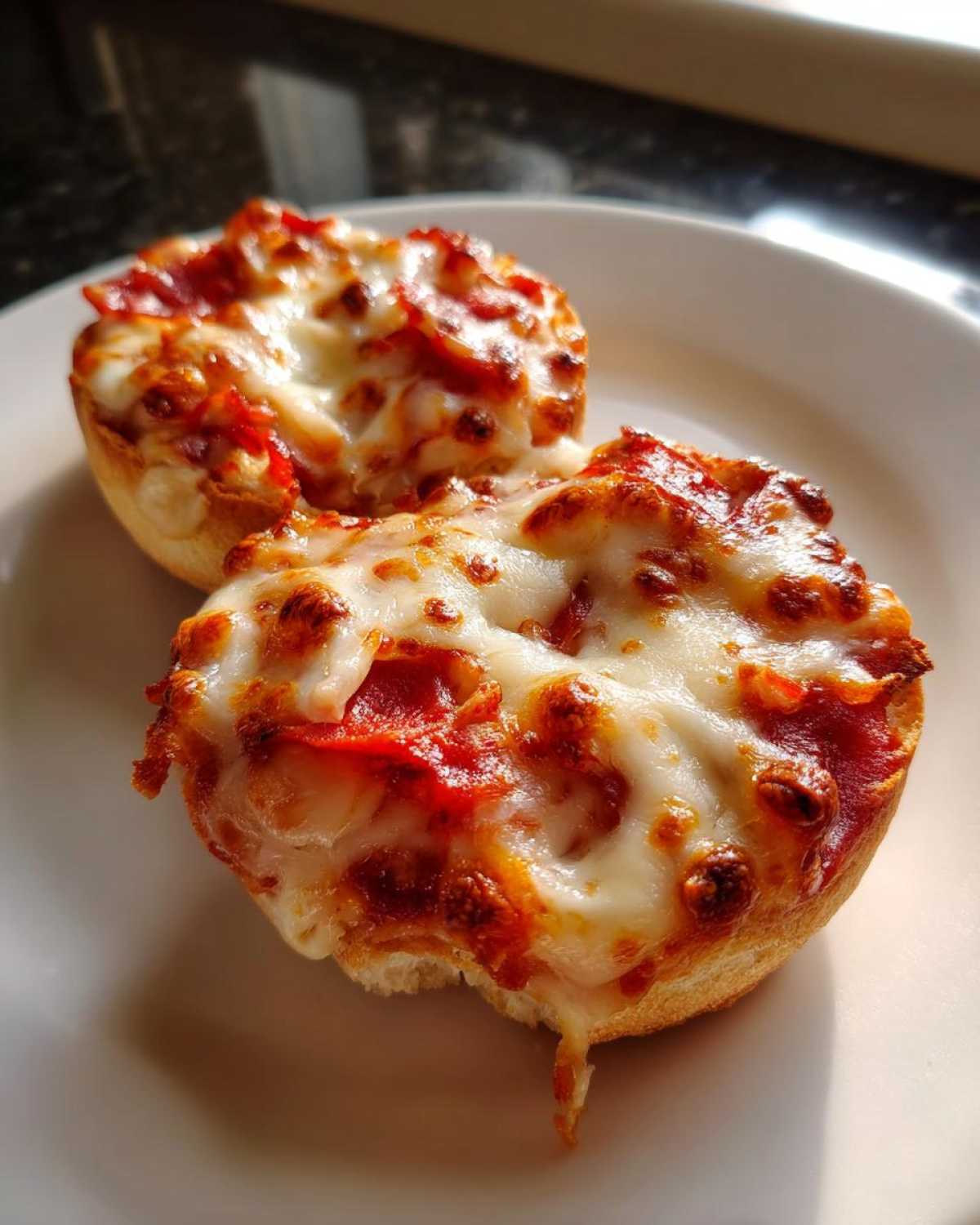 Two halves of a crispy English muffin pizza topped with melted, bubbly cheese and pepperoni, served on a white plate.