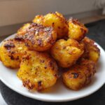 A close-up of a pile of golden, crispy Greek potatoes seasoned with herbs, served on a small white plate.