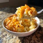 A spoonful of creamy crockpot cheesy potatoes, topped with melted, stringy cheddar cheese, being lifted from a white bowl.
