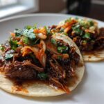 Two delicious Crockpot Korean Beef Tacos filled with shredded beef, kimchi, and cilantro on a white plate.