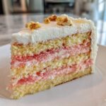 Close-up of a slice of Decadent Hawaiian Guava Cake showing yellow layers, pink guava filling, and white frosting topped with chopped nuts.