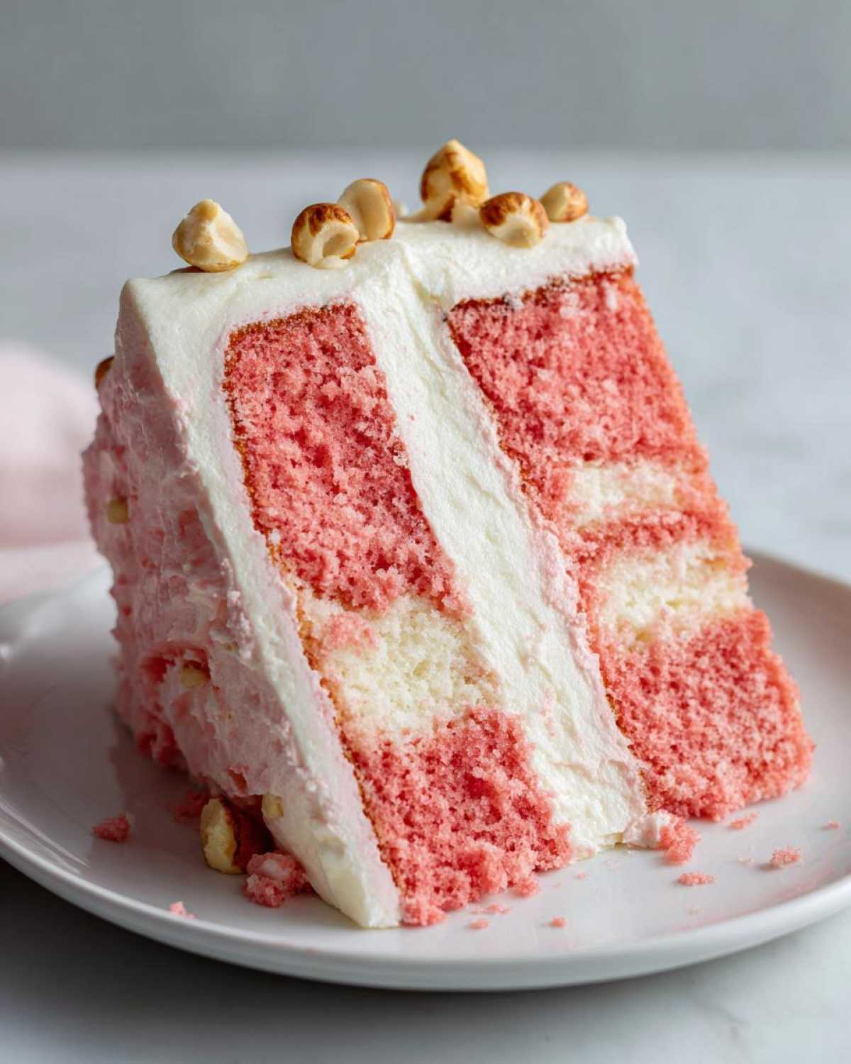 A tall slice of Decadent Hawaiian Guava Cake featuring pink and white layers, white frosting, and topped with chopped nuts.