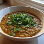 A close-up of a white bowl filled with rich, orange-brown Easy Green Lentil Dal, topped generously with fresh chopped cilantro.