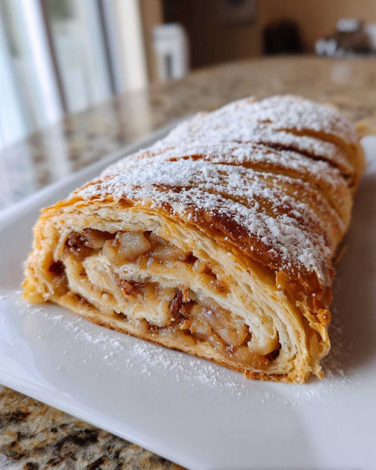 A cross-section view of a freshly baked apple strudel, dusted with powdered sugar, showing flaky pastry and apple filling.
