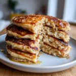 A tall stack of fluffy banana pancakes, with a bite taken out to show the texture, drizzled generously with syrup.