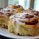 Close-up of several soft, freshly baked cinnamon buns covered in thick white icing on a white plate.