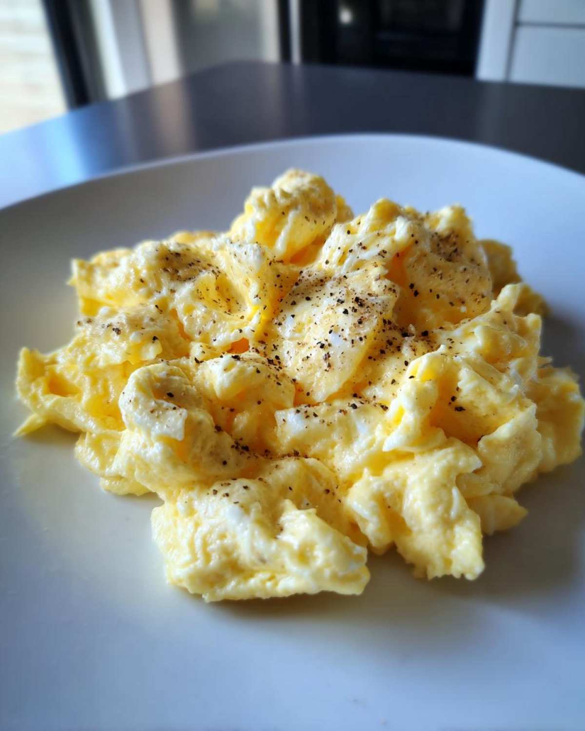 A close-up of creamy, fluffy scrambled eggs seasoned generously with cracked black pepper on a white plate.