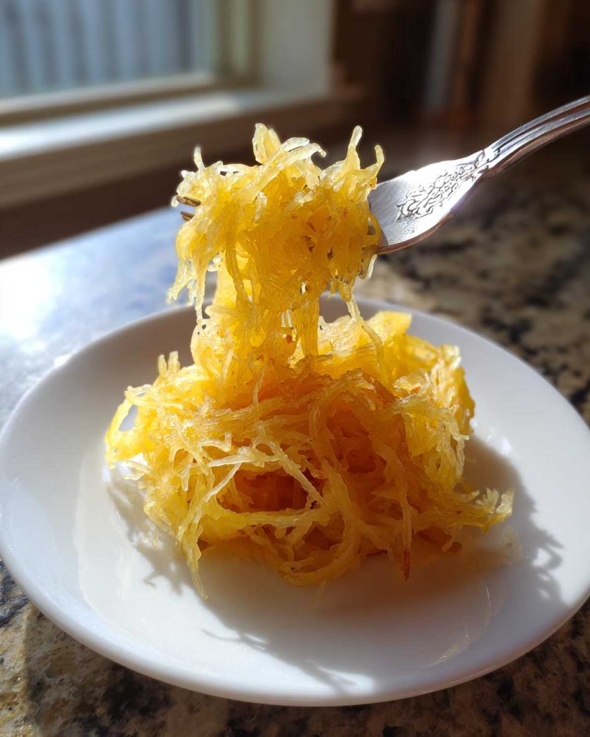 A fork lifts bright yellow, cooked strands of spaghetti squash recipe from a small white plate.