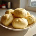 A pile of soft, pale yellow fufu balls served on a white plate near a sunlit window.