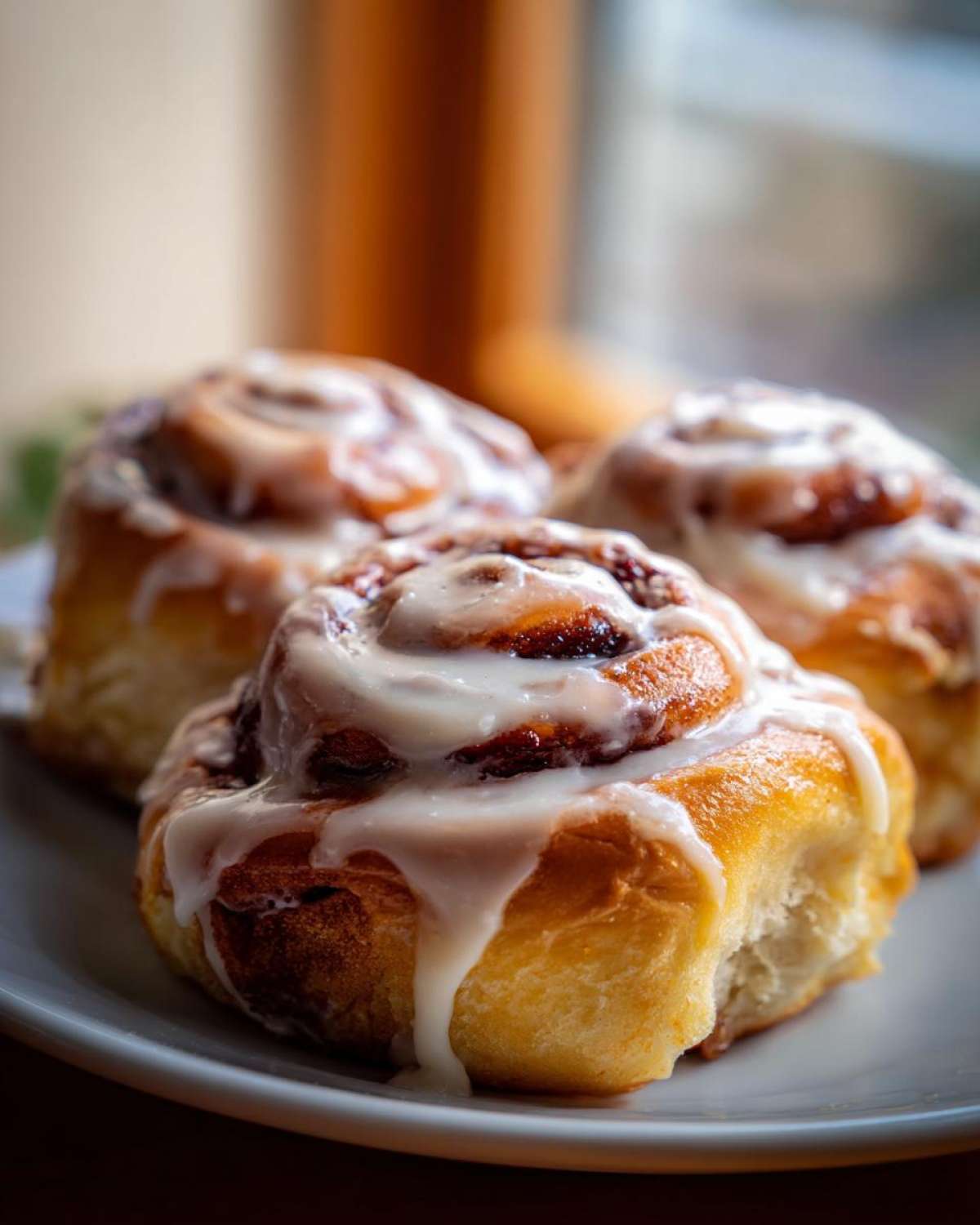 Three freshly baked, gooey cinnamon buns generously topped with white vanilla icing, sitting on a white plate.