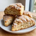 Four triangular lemon scones stacked on a white plate, drizzled with a white glaze and sprinkled with sugar.