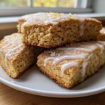 Close-up of several square lemon scones stacked on a white plate, drizzled with white glaze and sprinkled with lemon zest.