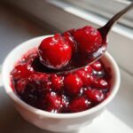 A spoonful of glossy, bright red cherry pie filling being lifted from a white bowl.