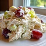 A close-up of a scoop of creamy greek yogurt chicken salad mixed with red and green grapes, served on a white plate.