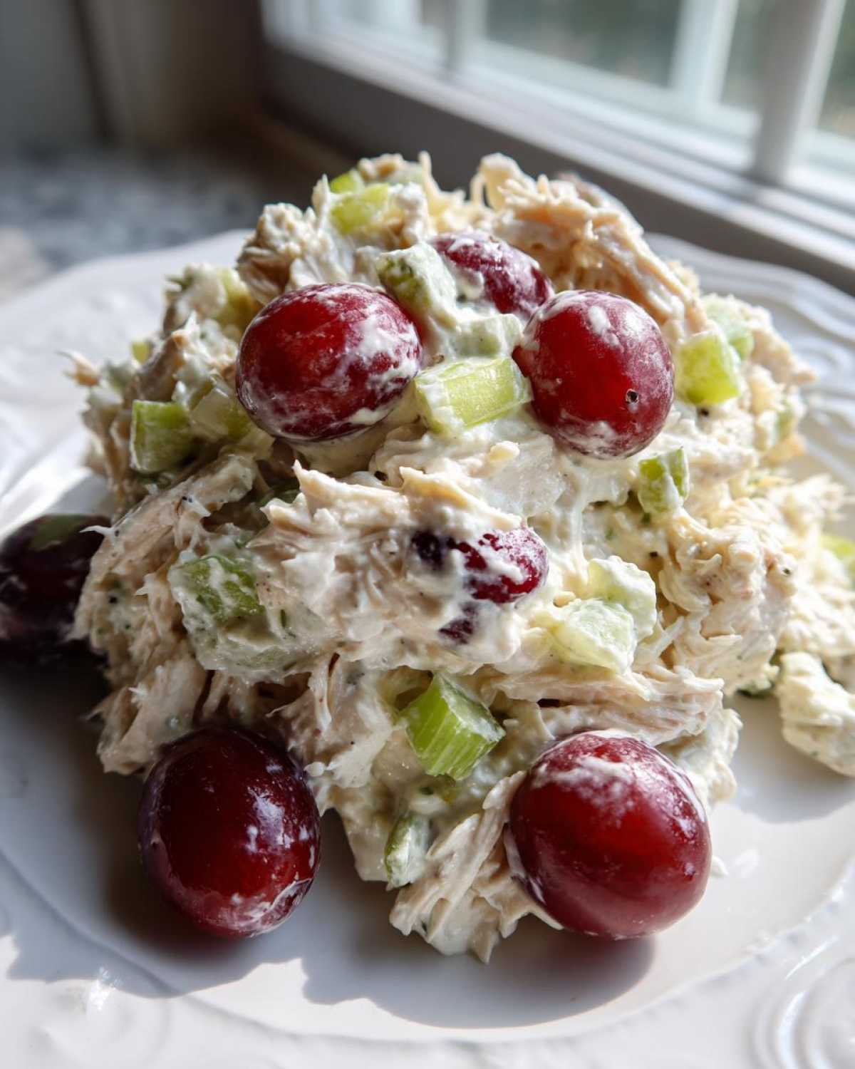 A scoop of creamy greek yogurt chicken salad mixed with celery and topped with whole red grapes on a white plate.