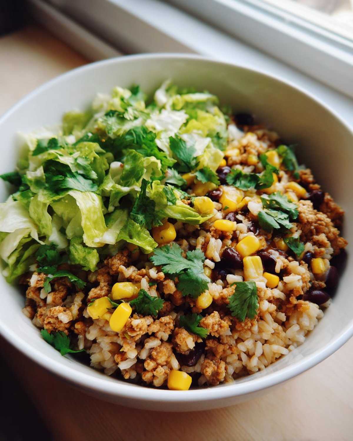 Close-up of a white bowl filled with seasoned ground turkey rice bowls, topped with corn, black beans, lettuce, and cilantro.