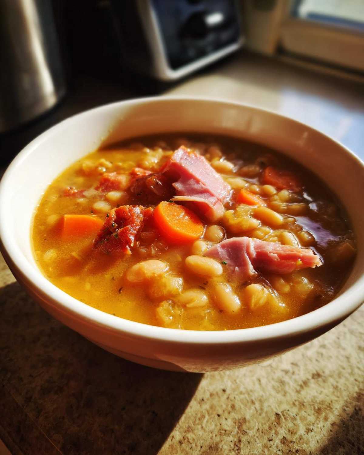 A close-up of a white bowl filled with thick navy bean soup, featuring chunks of ham and bright orange carrots.