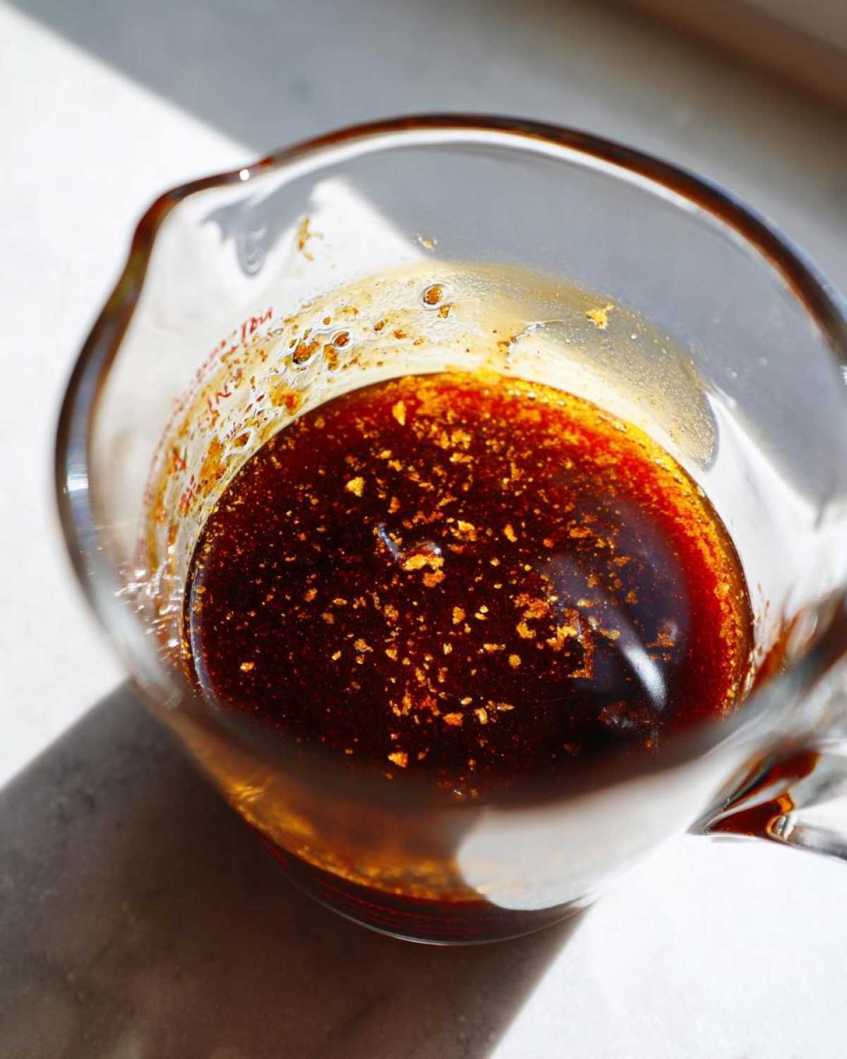 Close-up of rich, dark homemade teriyaki sauce with visible spices floating in a glass measuring cup.