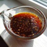 Close-up of rich, dark homemade teriyaki sauce with visible flecks of ginger or garlic in a small glass measuring cup.