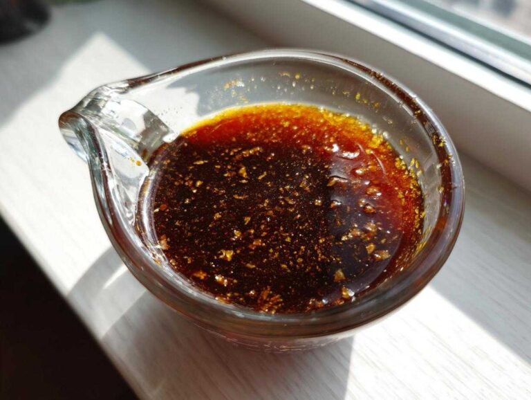 Close-up of rich, dark homemade teriyaki sauce with visible flecks of ginger or garlic in a small glass measuring cup.