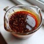 Close-up of rich, dark homemade teriyaki sauce with visible sesame seeds or spices floating on top, held in a glass measuring cup.