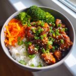 A close-up of a Honey Lime Chicken Rice Bowl featuring glazed chicken pieces over white rice, topped with scallions and sesame seeds, served with broccoli and shredded carrots.