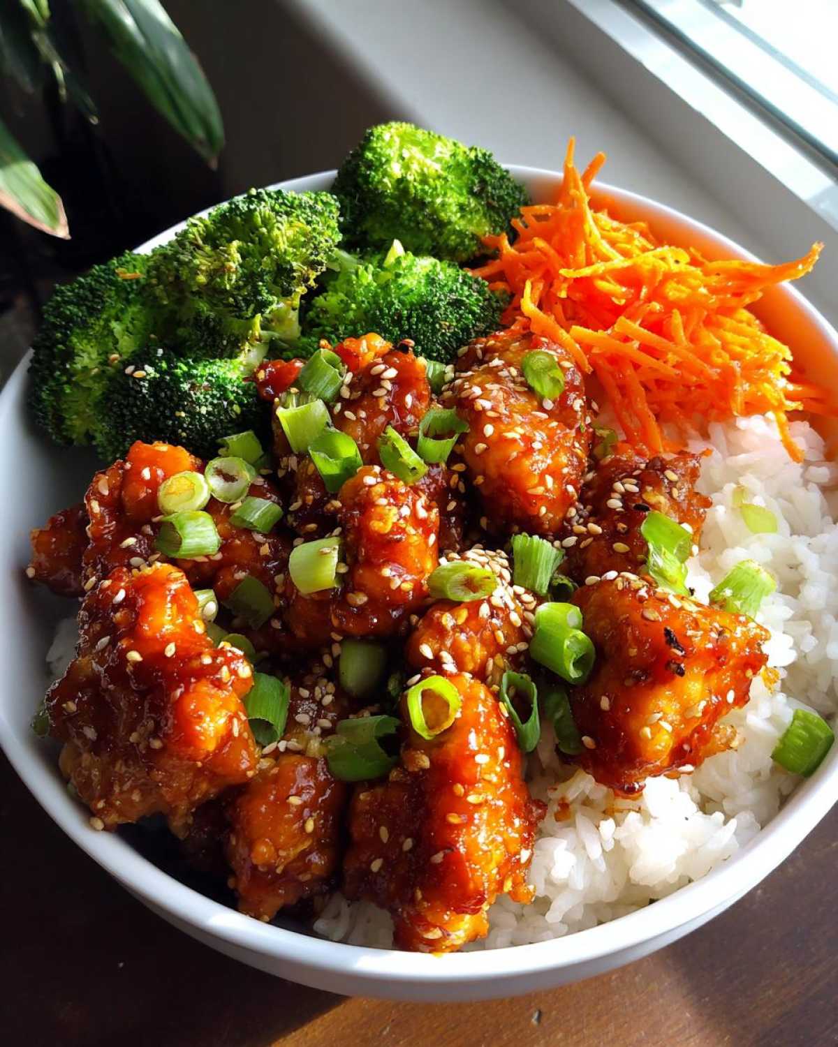 A close-up of a Honey Lime Chicken Rice Bowl featuring glazed chicken pieces over white rice, topped with sesame seeds, green onions, broccoli, and shredded carrots.