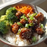 A close-up of a Honey Lime Chicken Rice Bowl featuring glazed chicken pieces over white rice, topped with sesame seeds, green onions, steamed broccoli, and shredded carrots.