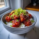 Close-up of a bowl filled with white rice topped with sticky, glazed Honey Sriracha Chicken and green onions.