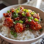 Close-up of a white bowl filled with white rice topped with glazed Honey Sriracha Chicken, sesame seeds, and chopped green onions.