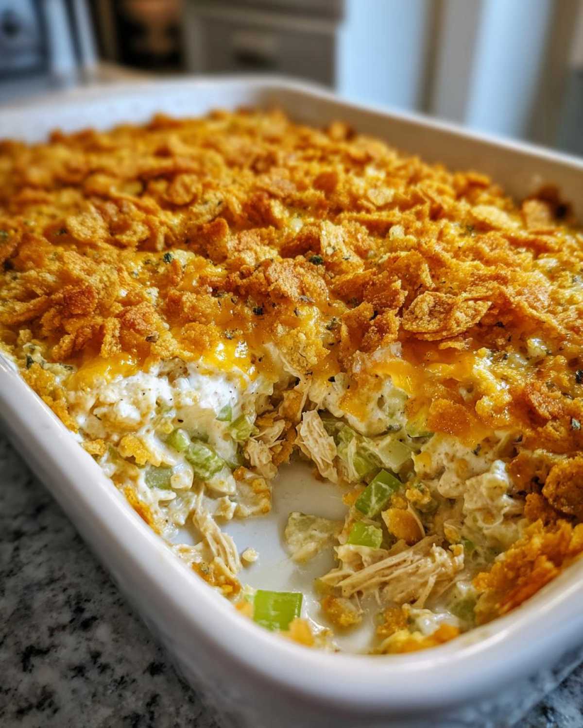 A close-up of a creamy hot chicken salad casserole in a white dish, featuring shredded chicken, celery, and a golden, crunchy cornflake topping.