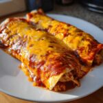 Two rolled Joanna Gaines Enchiladas smothered in red sauce and topped with melted cheddar cheese on a white plate.