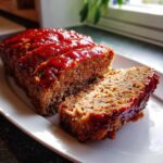 Close-up of a juicy meatloaf, sliced and topped with a thick, shiny red glaze, served on a white platter.