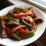 Close-up of a white plate filled with Keto Beef Pepper Steak strips, green peppers, red peppers, and onions in a savory sauce.