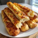 A stack of golden brown Keto Garlic Breadsticks, sprinkled with herbs, with one broken open showing a soft interior.