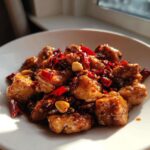 A close-up image of glistening, saucy Keto Kung Pao Chicken pieces mixed with dried red chilies and peanuts in a white bowl.