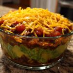 Close-up of a layered Avocado Party Dip featuring visible layers of green avocado, bean chili, and topped with shredded cheddar cheese.
