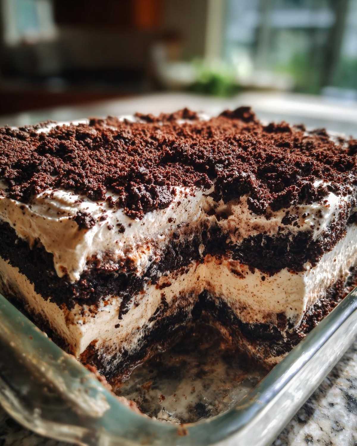 Close-up of a square glass dish showing a slice removed from the layered dirt cake, featuring alternating layers of cream and crushed chocolate cookies.