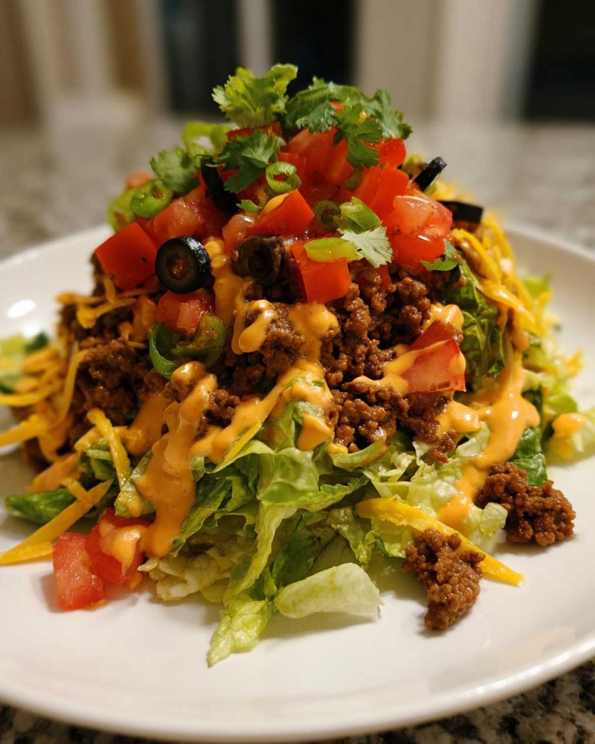 A tall, loaded portion of taco salad featuring seasoned ground beef, lettuce, cheese, tomatoes, olives, and creamy dressing.