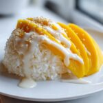 A mound of sweet sticky rice topped with sliced fresh mango, drizzled with coconut cream, and sprinkled with sesame seeds, representing mango sticky rice.