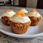 Four moist carrot cake muffins topped with thick cream cheese frosting and shredded carrots.