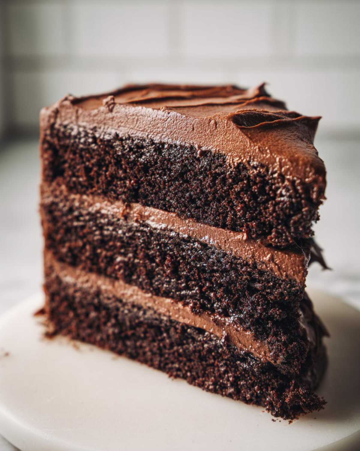 Close-up of a tall, moist slice of three-layer chocolate cake generously covered in dark chocolate frosting.