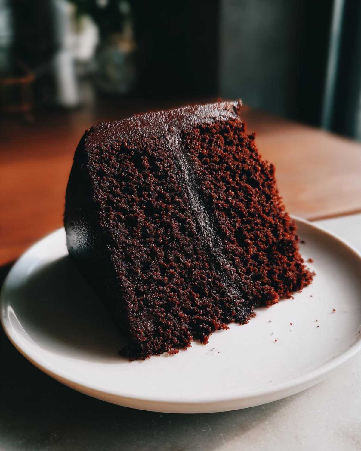 A rich, dark slice of moist chocolate cake topped with glossy chocolate frosting on a white plate.